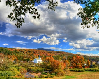 May include: A white church with a steeple stands on a hill overlooking a valley with colorful autumn foliage. The sky is blue with white clouds.