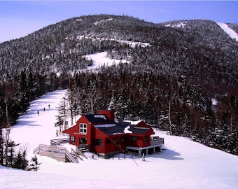 May include: A red wooden ski lodge with a snow-covered roof sits on a snowy hill with a forested mountain in the background.