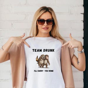 Puede incluir: Camiseta blanca con el texto "TEAM DRUNK" sobre una ilustración de dos monos de dibujos animados. Un mono sostiene una botella de cerveza. El texto "I'LL CARRY YOU HOME" está debajo.