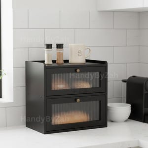 May include: A black two-drawer cabinet with glass-paneled doors and brass knobs. The cabinet sits on a white countertop, with a coffee mug, salt and pepper shakers, and other kitchen items on top. The cabinet has the text "HandyByZah" on the front.