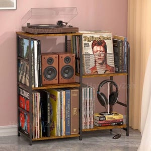 May include: A multi-tiered, rustic-style shelving unit with a record player, speakers, and vinyl records. The unit has a dark metal frame and wooden shelves. A Rolling Stone magazine featuring David Bowie is visible.