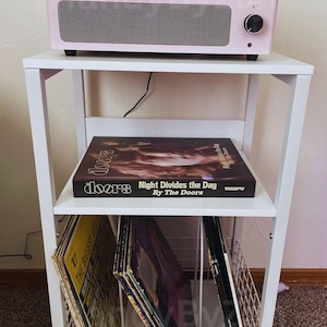 May include: A pink record player with a clear cover sits atop a white shelf unit. The shelf holds vinyl records, including a Doors album titled "Night Divides the Day." The unit is on a carpeted floor.