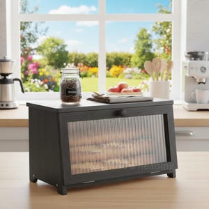 May include: A dark gray wooden bread box with a clear, ribbed glass door. A jar of basil sits on top, along with a plate of apples. The box is on a wooden countertop, with a window and a garden in the background.