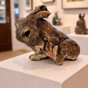 May include: A brown ceramic sculpture of a rabbit in a crouching position. The sculpture has a rough, textured surface with visible details. The rabbit's paws are a light pink colour. The sculpture is displayed on a white plinth.