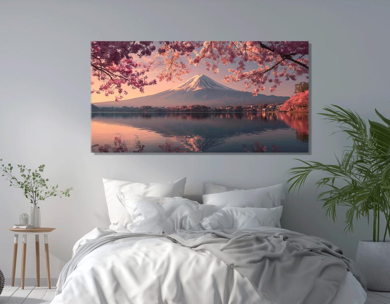 May include: A canvas print of a serene landscape featuring a snow-capped mountain reflected in calm water. Pink cherry blossoms frame the scene, with a soft, warm color palette. The artwork is displayed above a bed with white linens.