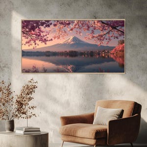 May include: A framed landscape print of Mount Fuji, Japan, reflected in a lake. The image features a snow-capped mountain, pink cherry blossoms, and a serene, mirrored water surface. The print is displayed on a textured gray wall, with a brown armchair and side table in the foreground.