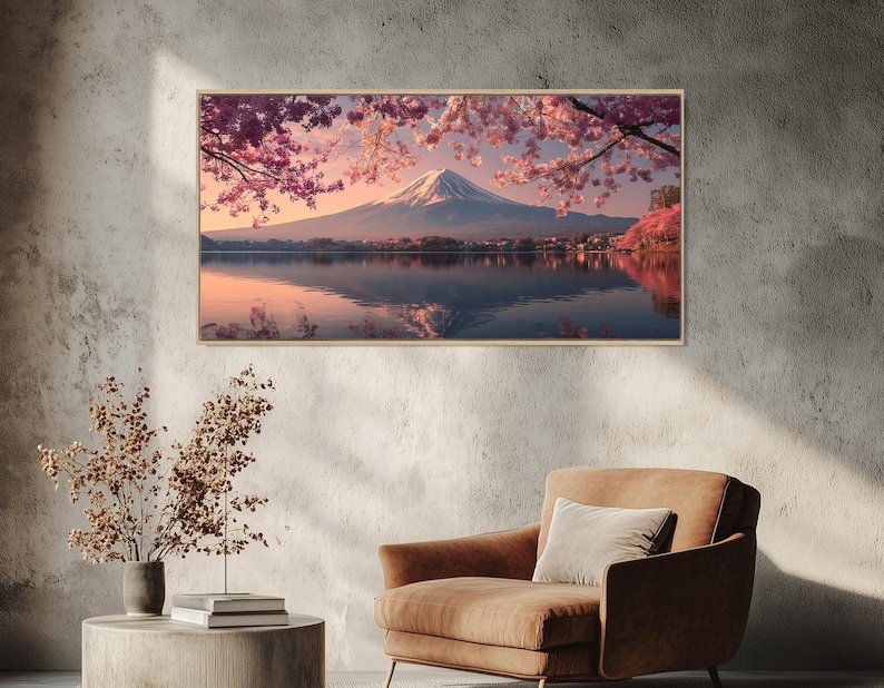 May include: A framed landscape print of Mount Fuji, Japan, reflected in a lake. The image features a snow-capped mountain, pink cherry blossoms, and a serene, mirrored water surface. The print is displayed on a textured gray wall, with a brown armchair and side table in the foreground.