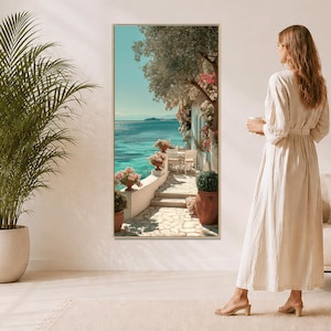 May include: A framed vertical art print shows a coastal scene with a white stone pathway, turquoise water, and flowering plants. The artwork is in a light-coloured frame. A woman in a long dress stands nearby, holding a cup.