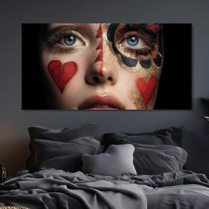 Könnte beinhalten: Nahaufnahme eines Leinwanddrucks mit einem Gesicht mit auffälligem Make-up. Das Make-up umfasst rote Herzen, goldene Akzente und schwarze Details. Die Person hat blaue Augen und rote Lippen. Der Hintergrund ist schwarz.
