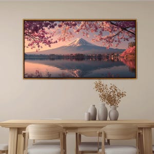 May include: A framed landscape print featuring Mount Fuji, Japan, reflected in a lake. The image showcases cherry blossoms in full bloom, with pink and white petals framing the mountain. The artwork is displayed above a wooden table.