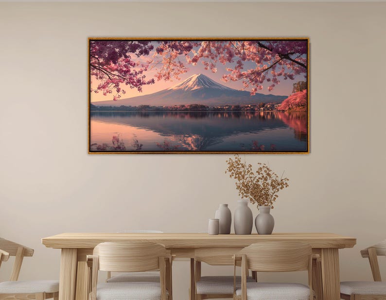 May include: A framed landscape print featuring Mount Fuji, Japan, reflected in a lake. The image showcases cherry blossoms in full bloom, with pink and white petals framing the mountain. The artwork is displayed above a wooden table.