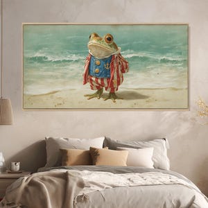 Puede incluir: Una impresión artística enmarcada representa una rana vestida con un traje de marinero en una playa. La rana lleva una chaqueta azul con botones de ancla y pantalones a rayas rojas y blancas. El fondo muestra un mar turquesa y una playa arenosa.