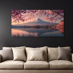 May include: A canvas print depicting a serene landscape. The image showcases a snow-capped mountain reflected in a calm lake, framed by pink cherry blossoms. The artwork is displayed above a beige sofa with several pillows.