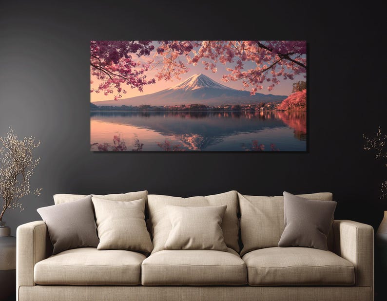 May include: A canvas print depicting a serene landscape. The image showcases a snow-capped mountain reflected in a calm lake, framed by pink cherry blossoms. The artwork is displayed above a beige sofa with several pillows.