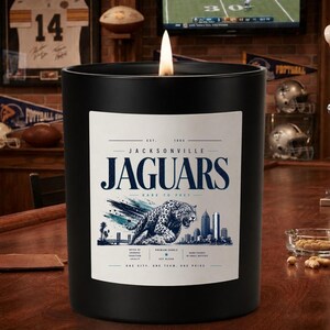 May include: A black cylindrical candle with a white label featuring the text Jacksonville Jaguars, a graphic of a leaping jaguar, and a city skyline. The label includes the text Est. 1995, Dare to Prey, and One City, One Team, One Pride.
