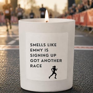 May include: A white candle in a cylindrical container with a lit wick. The label features the black text: SMELLS LIKE EMMY IS SIGNING UP GOT ANOTHER RACE, accompanied by a small black silhouette graphic of a person running.