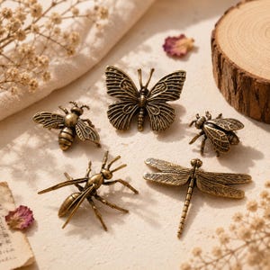 Könnte beinhalten: Eine Sammlung antiker Insektenfiguren aus Messing, darunter ein Schmetterling, eine Biene, eine Libelle und zwei weitere Insekten. Die detaillierte Metallarbeit zeigt die Flügel und Körpersegmente jedes Insekts. Die Figuren sind auf einer neutralen Oberfläche angeordnet.
