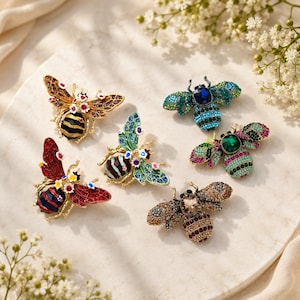 Pode incluir: Uma coleção de seis broches em forma de inseto com trabalhos em metal intrincados e adornos de cristais coloridos. Cada broche exibe padrões únicos, incluindo detalhes florais, corpos listrados e centros de pedras preciosas em vários tons.