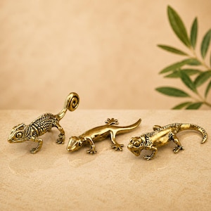 May include: Three small, detailed brass lizard figurines. One has a curled tail, another has a straight tail, and the third has a short tail. The lizards are posed on a light-coloured surface with a neutral background.