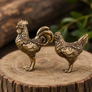 Peut inclure: Deux figurines en métal de couleur laiton représentant un coq et une poule. Le coq se tient à gauche avec une crête proéminente et des plumes de queue détaillées, tandis que la poule plus petite se tient à droite. Plumage texturé.