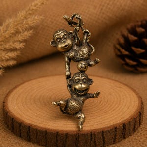 Könnte beinhalten: Eine messingfarbene Metallfigur, die zwei miteinander verbundene Affen darstellt. Der obere Affe hängt an einem gebogenen Haken, während der untere Affe eine kleine Kugel hält, in die das chinesische Schriftzeichen für Reichtum eingraviert ist.