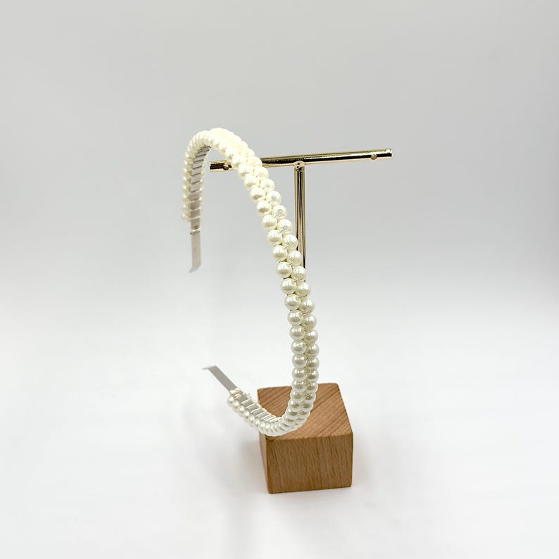 May include: A cream-coloured headband featuring two rows of faux pearls. The headband is displayed on a gold-coloured metal stand with a wooden base. The background is plain white.