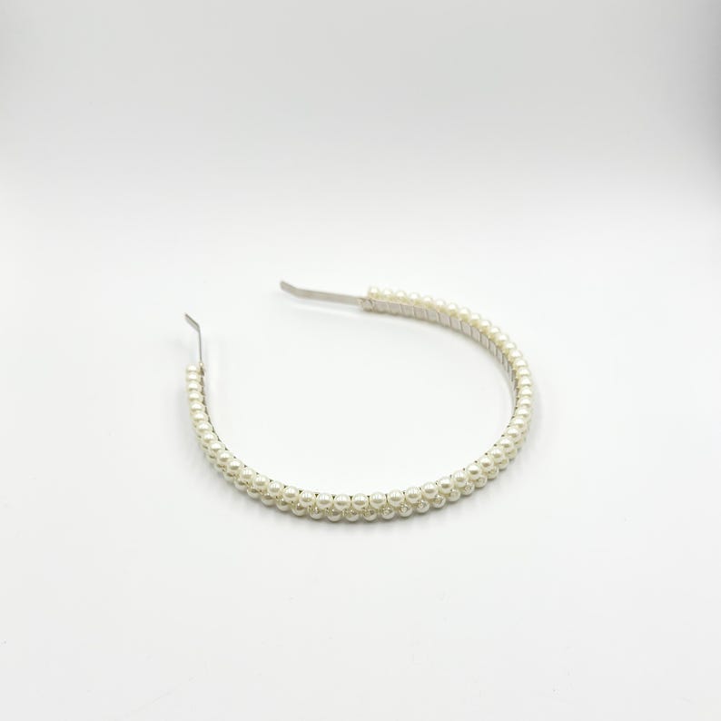 May include: A white headband adorned with small, round, off-white pearls. The headband features a silver-coloured metal piece at each end. The pearls are evenly spaced, forming a continuous line around the band.