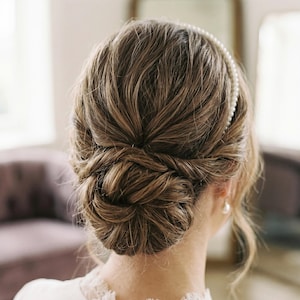 May include: A woman's brown hair styled in an updo, viewed from the back. A pearl headband and earring are visible. The hairstyle is intricate and elegant, suitable for a wedding or formal event. The hair is styled in a bun.
