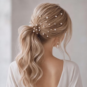 Op de afbeelding: Een blond paardenstaartkapsel versierd met talloze kleine, ronde witte parelhaarspelden die door het haar verspreid zijn. Het haar is gestyled in een losse, golvende paardenstaart met lokken die het gezicht omlijsten en een zichtbare pareloorbel.