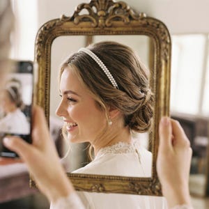 May include: A woman wearing a double-strand pearl headband and pearl earrings, framed by a gold ornate mirror. The woman is smiling, and her hair is styled in an updo. The image is taken indoors, with soft lighting.