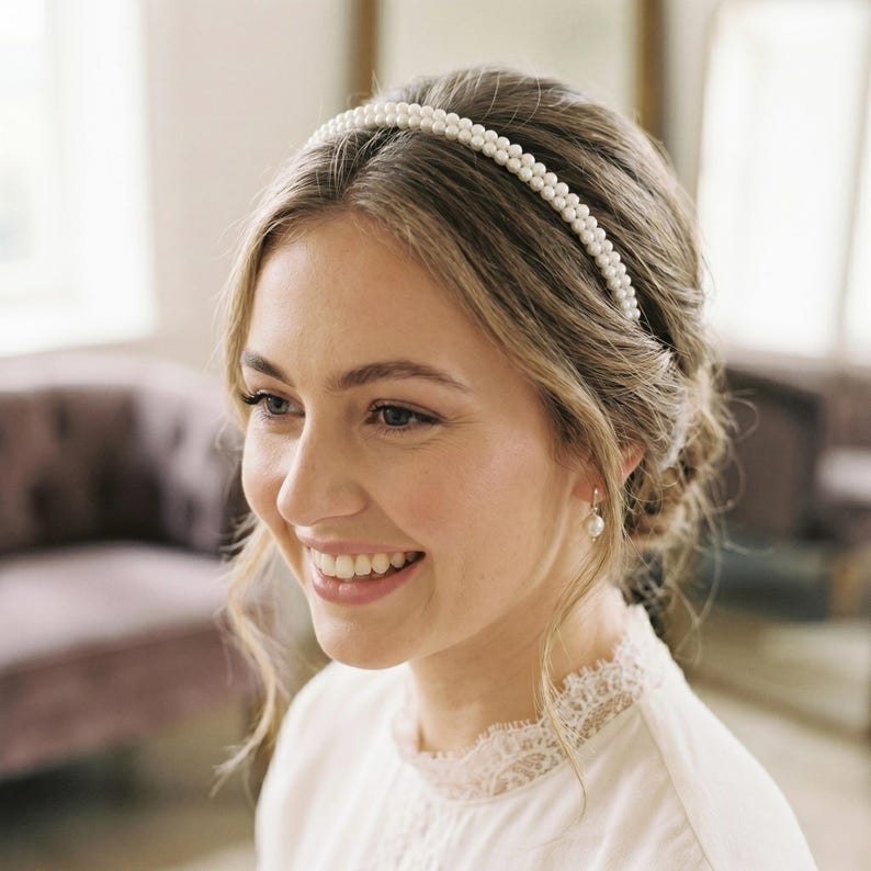 May include: A double-strand pearl headband. The white pearls are arranged in a neat row. The woman is wearing a white blouse with lace trim and pearl earrings. The background is softly lit with a blurred interior.