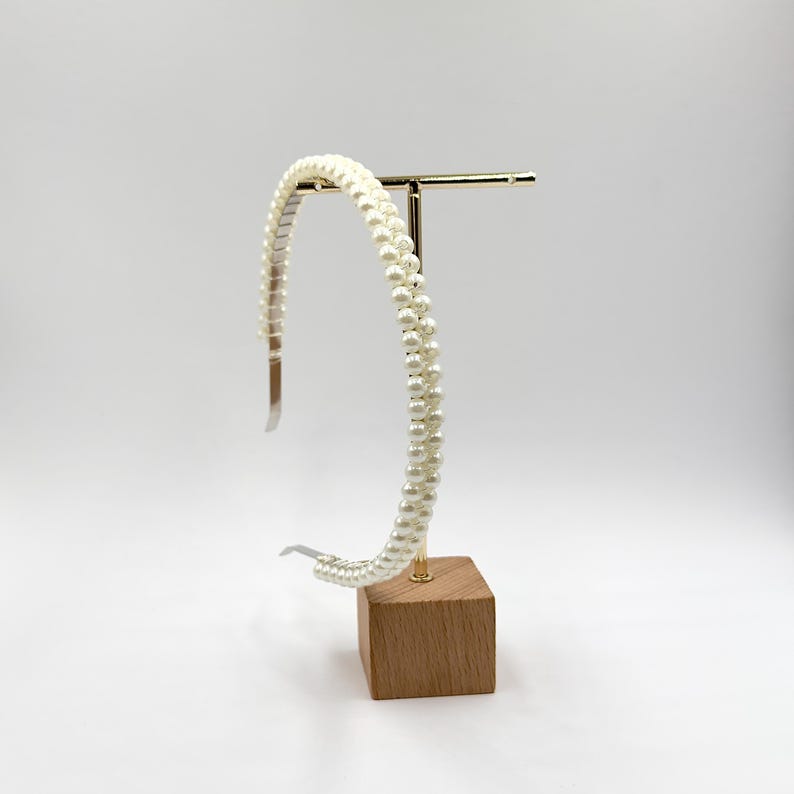 May include: A cream-coloured pearl headband with a double strand design. The headband is displayed on a gold-coloured metal stand with a small wooden base. The pearls are small and evenly spaced, creating a classic and elegant look.