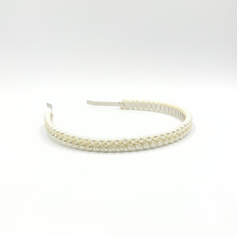May include: A white faux pearl headband. The headband has two rows of small, round, off-white pearls. The band is made of silver-coloured metal. The pearls are evenly spaced, creating a delicate and elegant appearance.
