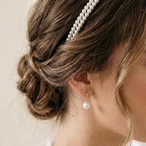 May include: A woman's hair styled in an updo, with a double-strand pearl headband. A single pearl earring is visible. The hair is light brown. The image is a close-up.