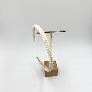 May include: A white pearl beaded headband. The headband is displayed on a gold-coloured metal stand with a wooden base. The pearls are arranged in two parallel rows, creating a textured, elegant look.