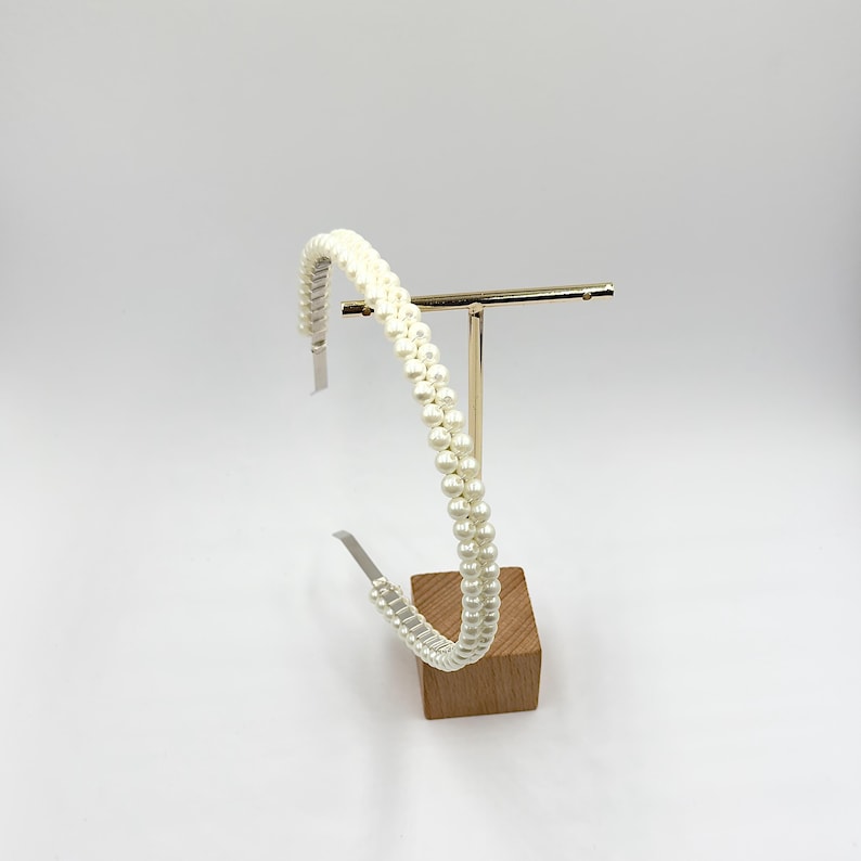 May include: A white pearl beaded headband. The headband is displayed on a gold-coloured metal stand with a wooden base. The pearls are arranged in two parallel rows, creating a textured, elegant look.