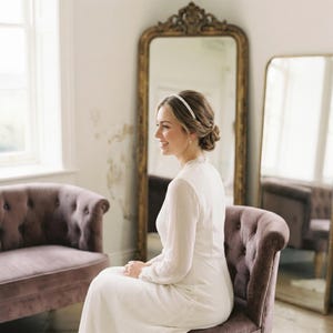 May include: A woman in a white dress and pearl headband sits on a purple velvet chair. A large gold-framed mirror is behind her. The room has a vintage aesthetic with a matching purple sofa and a window.