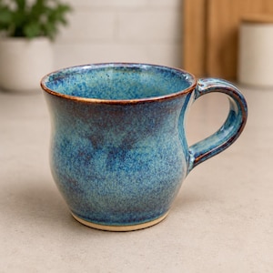 May include: A handmade ceramic mug with a blue glaze. The mug has a rounded body, a curved handle, and a brown rim. The blue glaze has a mottled appearance, with darker and lighter areas. The mug is sitting on a light-colored surface.