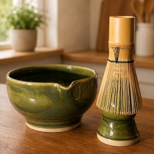 May include: A dark green ceramic bowl with a pouring spout and a bamboo matcha whisk resting on a matching green ceramic stand. The bowl features a smooth, glossy finish with subtle color variations, while the whisk has thin, light-colored tines.