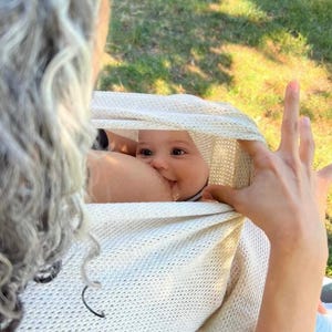 Op de afbeelding: Een lachende baby is genesteld in een crèmekleurige gebreide omslagdoek, met de arm en hand van een persoon zichtbaar. De baby drinkt borstvoeding, met een deel van de arm en hand van de persoon zichtbaar. De achtergrond is een vage buitenscène met groen gras.