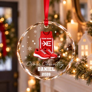 May include: A circular crystal ornament hanging from a red ribbon. The design features a red tank top with the text TEAM NAME and XE, a pair of red running shoes, and the text Cross Country, DANIEL, and 2026, surrounded by white snowflakes.