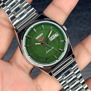 May include: A silver-tone stainless steel Seiko automatic wristwatch featuring a round green dial with a wavy pattern, silver hour markers, and a day-date window at the three o'clock position. The watch includes a matching silver metal link bracelet.