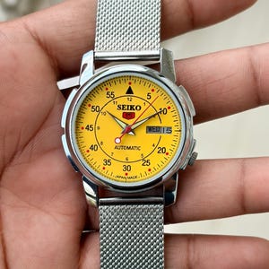 May include: A Seiko 5 automatic wristwatch with a bright yellow dial, silver-tone hands, and a day-date window at the three o'clock position. The watch features a stainless steel case and a silver-colored mesh metal strap with a secure clasp.