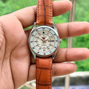 May include: A Seiko 5 automatic wristwatch with a cream-colored dial, featuring both 12-hour and 24-hour markers, a day-date window at the three o'clock position, and a brown leather strap with a textured, embossed crocodile-style pattern.
