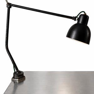 Könnte beinhalten: Eine schwarze Schreibtischlampe aus Metall mit verstellbarem Arm und kuppelförmigem Lampenschirm. Die Lampe verfügt über einen Klemmfuß zur Befestigung an einer Oberfläche und ein grün stoffummanteltes Kabel am Arm.