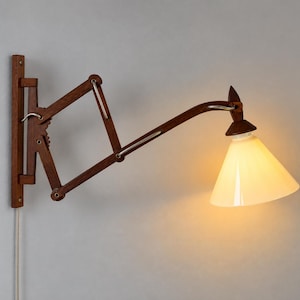 Könnte beinhalten: Eine Wandleuchte mit einem dunkelbraunen, scherenartigen Ziehharmonika-Arm aus Holz und einem konischen weißen Glasschirm. Die Lampe leuchtet warm und verfügt über eine vertikale Wandhalterung aus Holz sowie ein weißes Stromkabel.