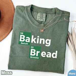 Puede incluir: Una camiseta verde musgo con las palabras "Baking Bread" en un diseño de estilo de tabla periódica. La camiseta también tiene la palabra "Moss" en la parte inferior izquierda. Un collar dorado está drapeado sobre la camiseta.