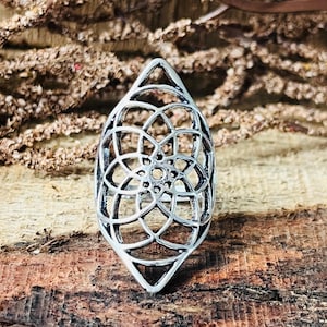 Könnte beinhalten: Ein silberfarbener Ring mit einem durchbrochenen Design. Der Ring weist ein geometrisches Muster aus miteinander verbundenen Formen auf, das ein blumen- oder mandalaartiges Aussehen erzeugt. Der Ring ist vor einem Holzgrund platziert.