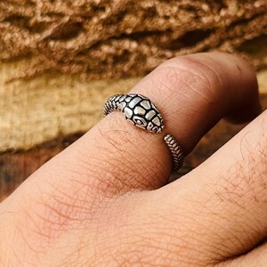May include: A silver snake ring with a detailed head and textured body. The ring is open-ended, allowing for adjustable sizing. The snake's head features a scale pattern, and the eyes are visible.