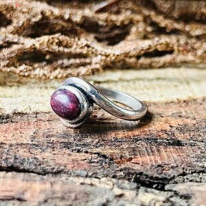 May include: A silver ring featuring a dark red oval gemstone. The gemstone is set in a bezel setting, and the band has a swirling design. The ring is displayed on a textured wooden surface.